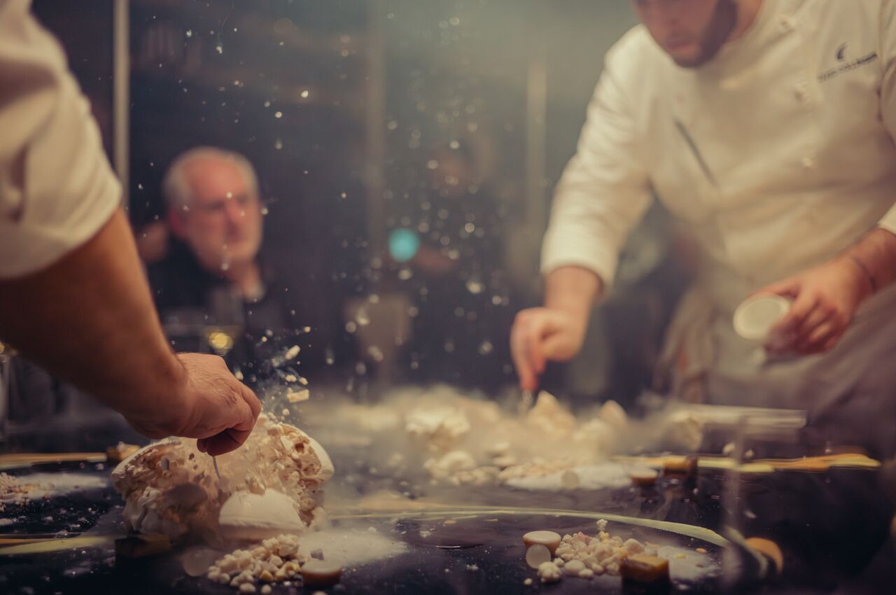 Two chefs in white uniforms create clouds of powdered sugar and meringue while preparing a dessert at a restaurant table.