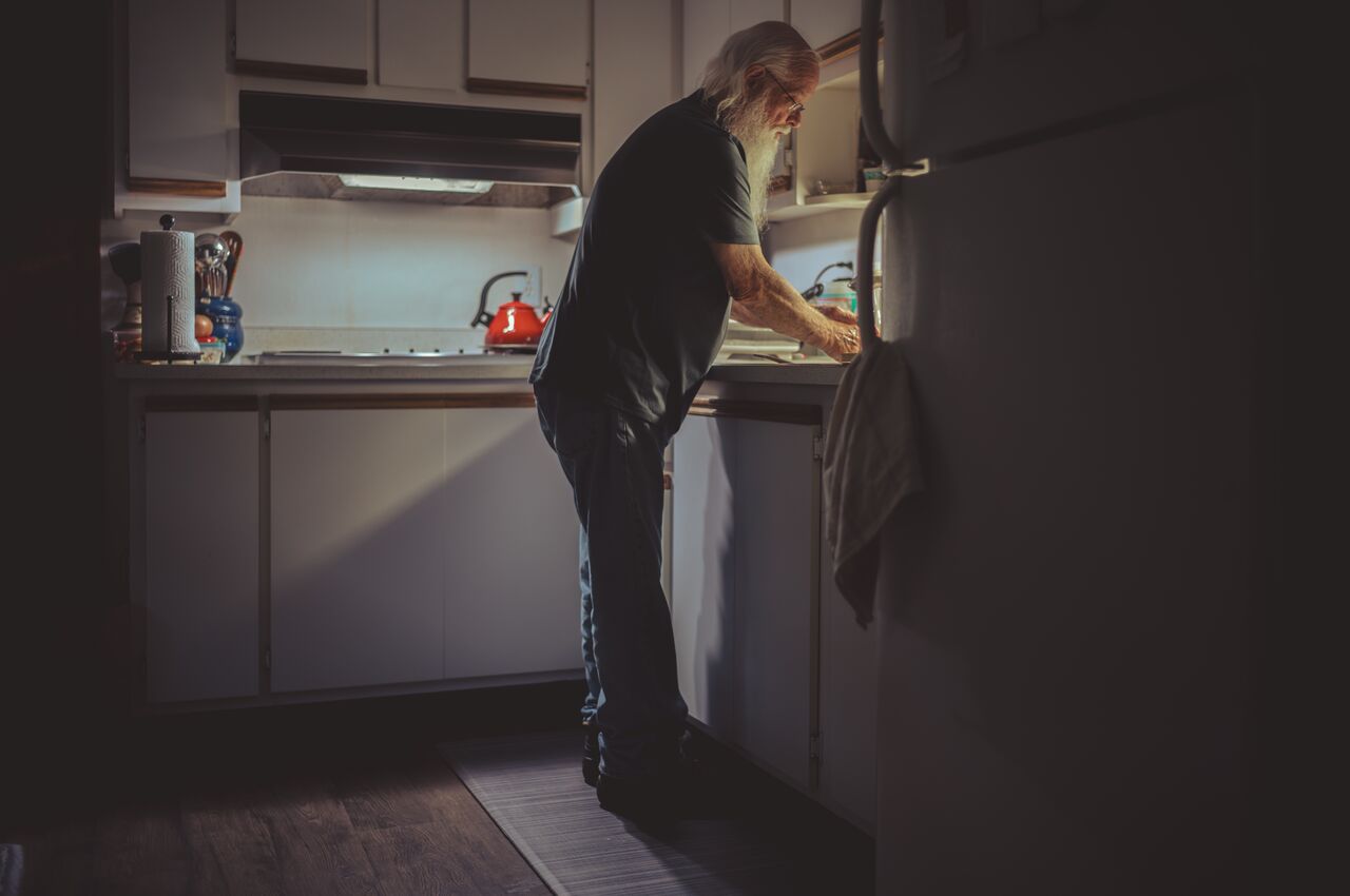 An older man stands at the sink, doing the dishes by hand under a warm light, while the kitchen around him is dark and quiet.