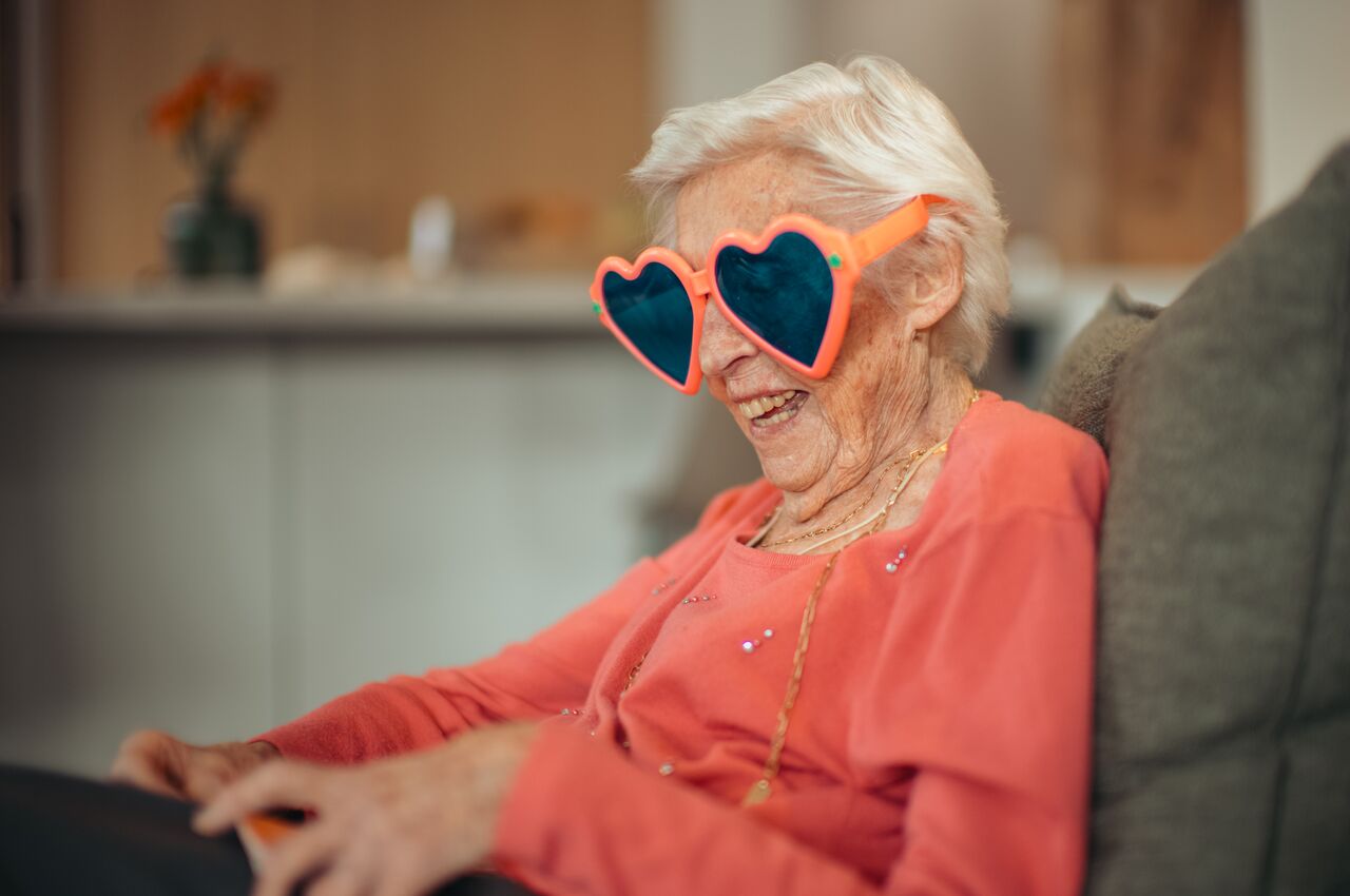 An elderly woman with white hair wearing pink top and heart-shaped sunglasses, laughing while seated indoors.