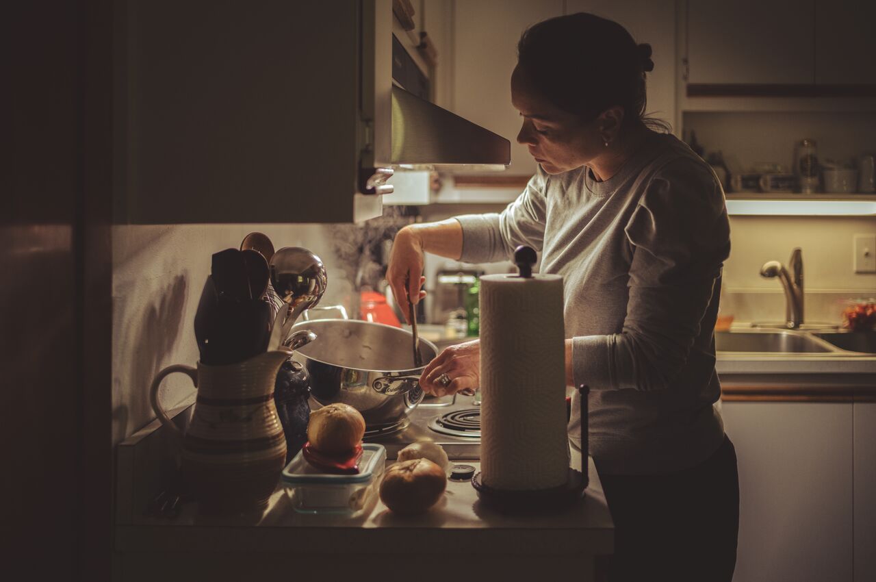 A woman stands at the stove, stirring a pot and cooking dinner in a warm, softly lit kitchen.