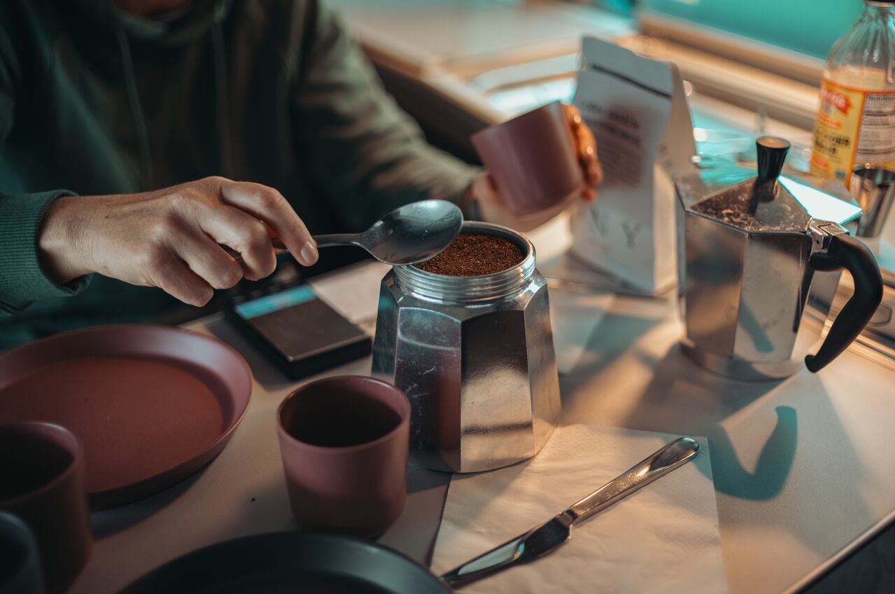 A person scooping ground coffee into the filter basket of a moka pot.