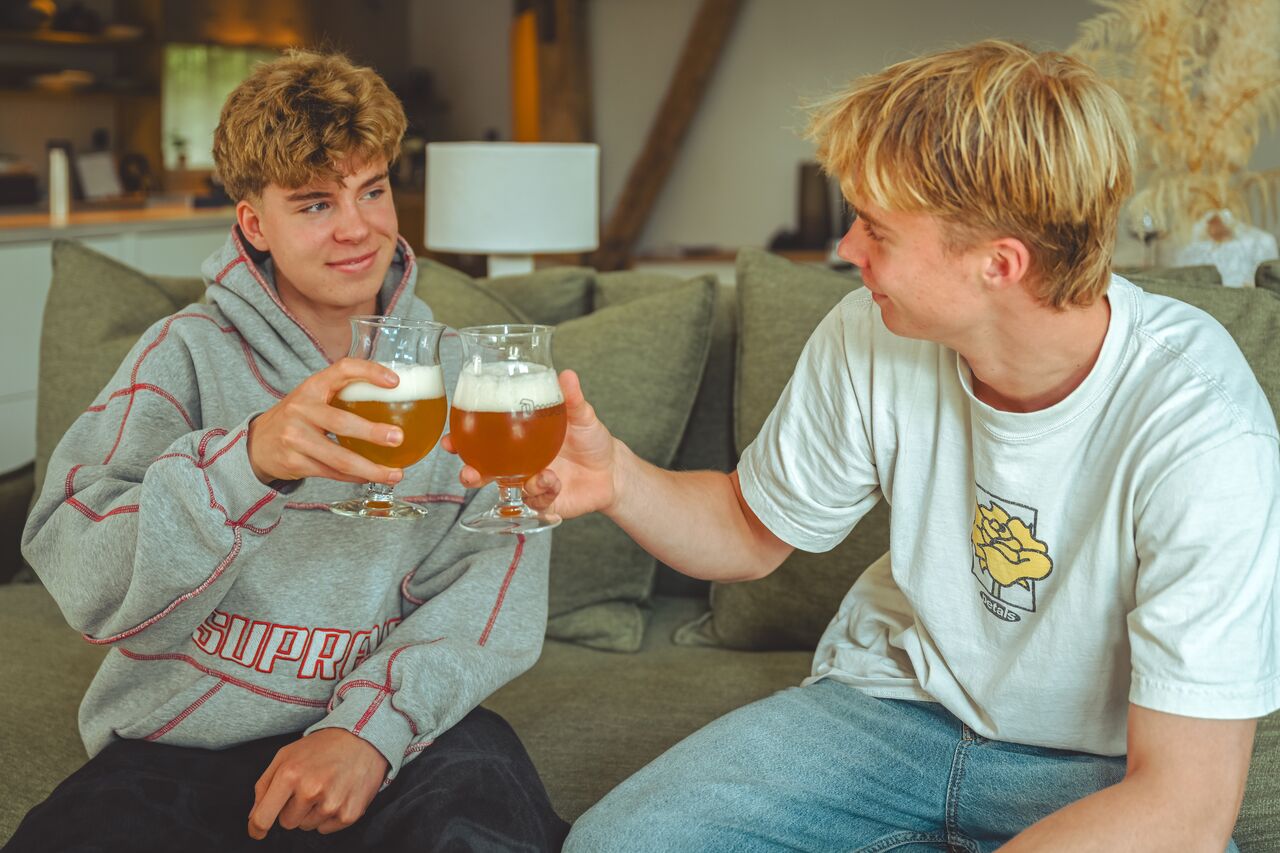 Two young adults sit on a couch, smiling at each other as they clink glasses of beer.
