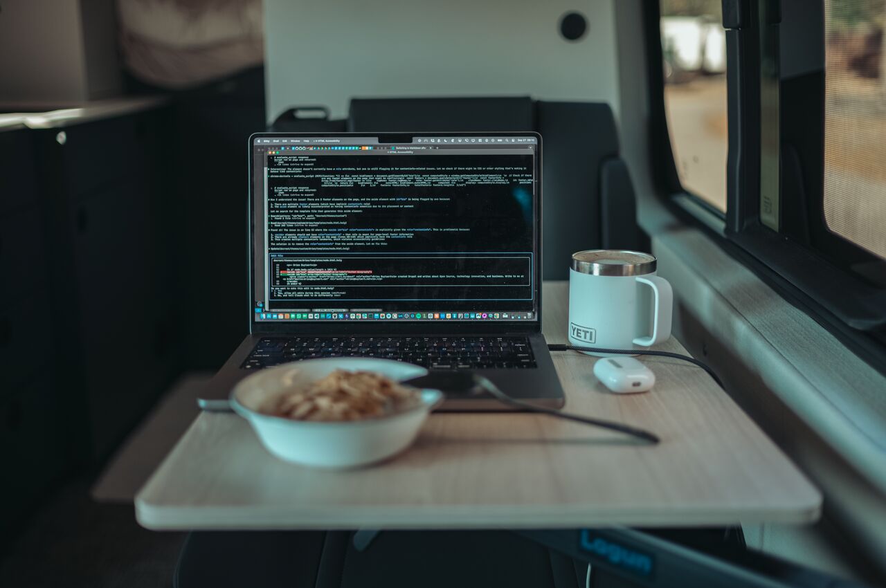 A laptop open on a small table inside a van, showing code on the screen, with a bowl of cereal and a mug of coffee beside it.