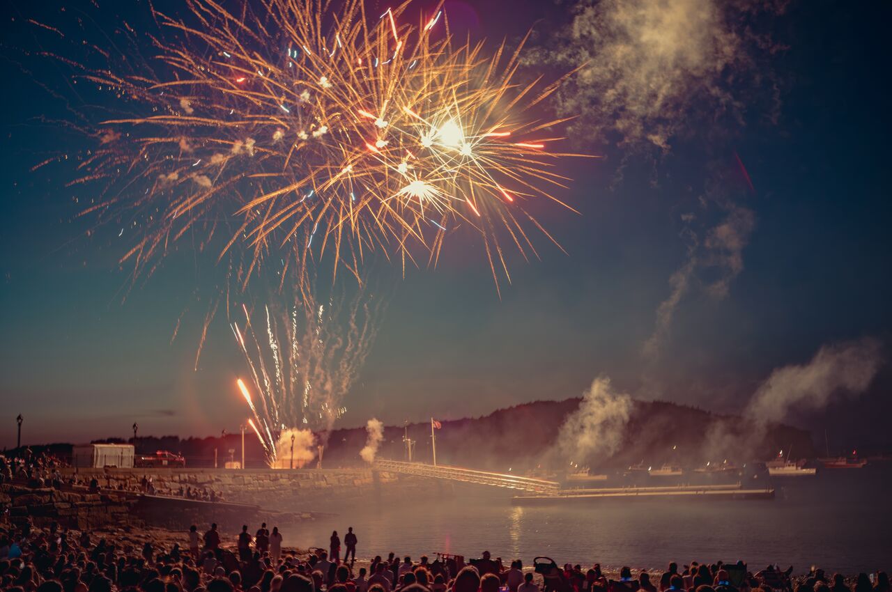 Alt text: Fireworks over the water in Bar Harbor, Maine, with boats in the harbor and a packed beach watching from below.