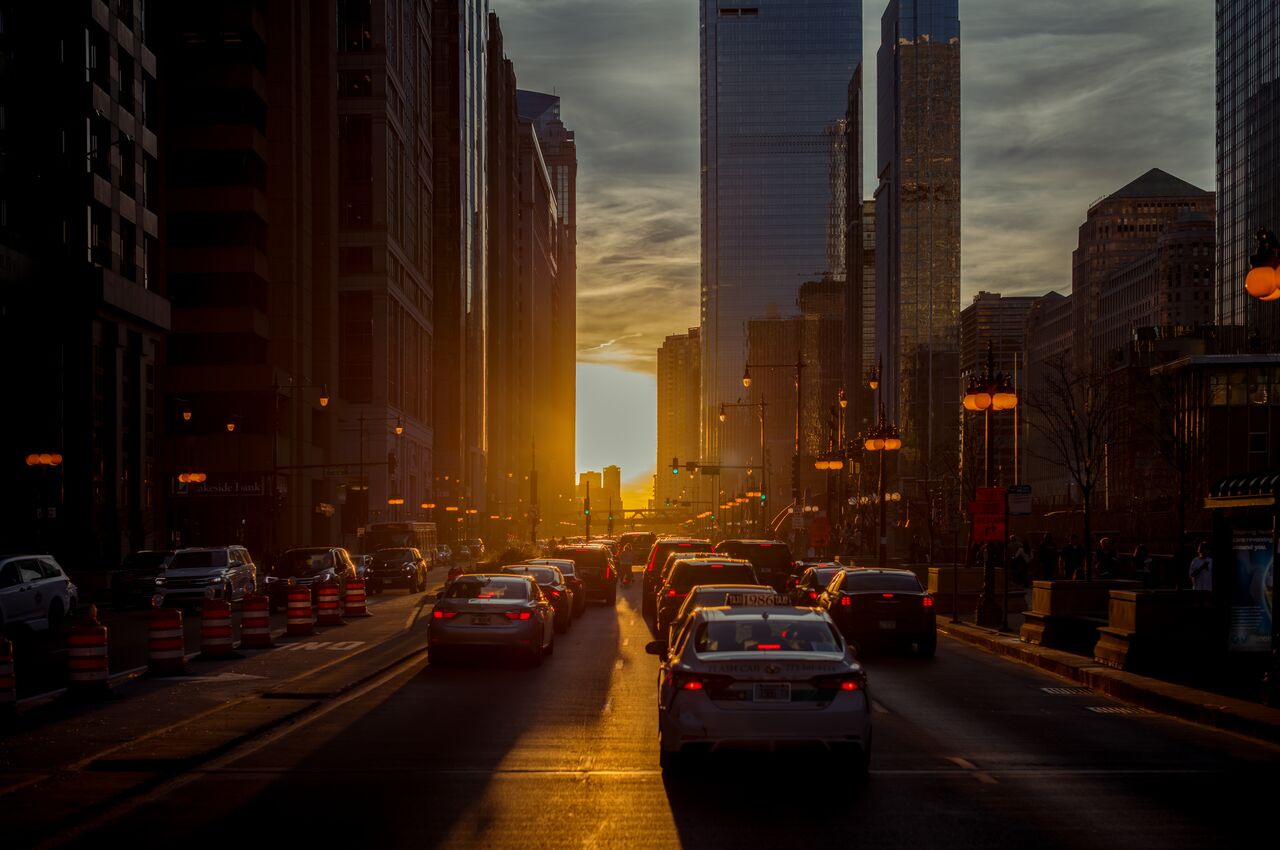 Cars line up on a wide street in downtown Chicago, with tall buildings on both sides and the sun low between them.