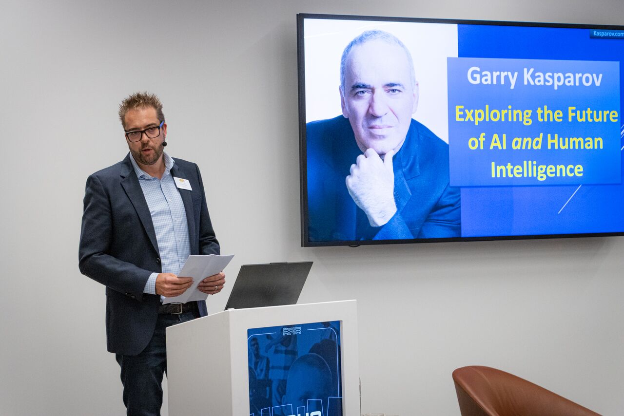 A man in a blazer and glasses stands at a lectern holding papers, beside a screen showing Garry Kasparov and the title "Exploring the Future of AI and Human Intelligence".