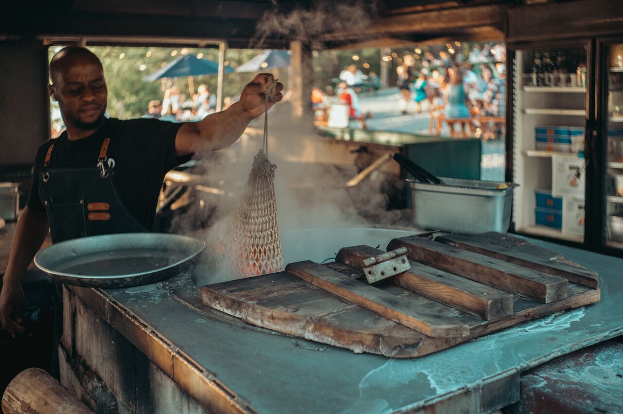 A cook lifts a mesh bag of freshly steamed lobster from a steaming kettle.