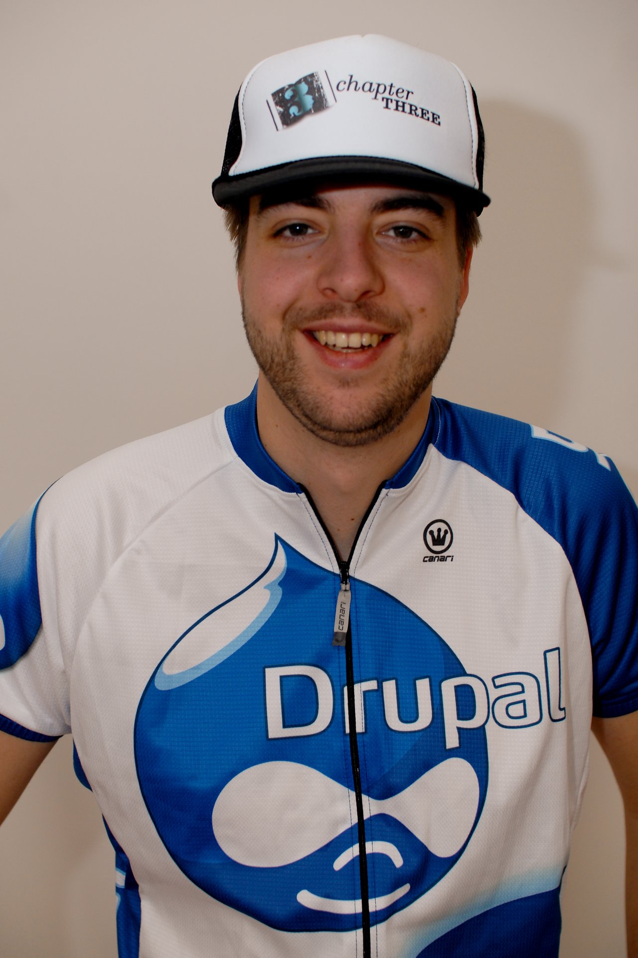 A smiling person wears a Drupal cycling jersey and a cap with "Chapter Three" printed on it.