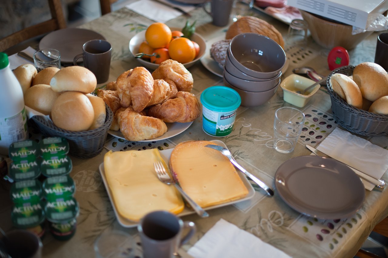 A breakfast table with croissants, bread rolls, cheese, yogurt, fruit, and drinks, set for multiple people.