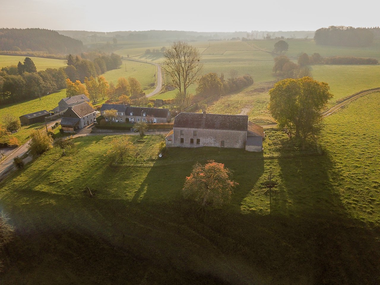 Aerial view of a rural farmhouse and surrounding green fields in the Ardennes during late afternoon.