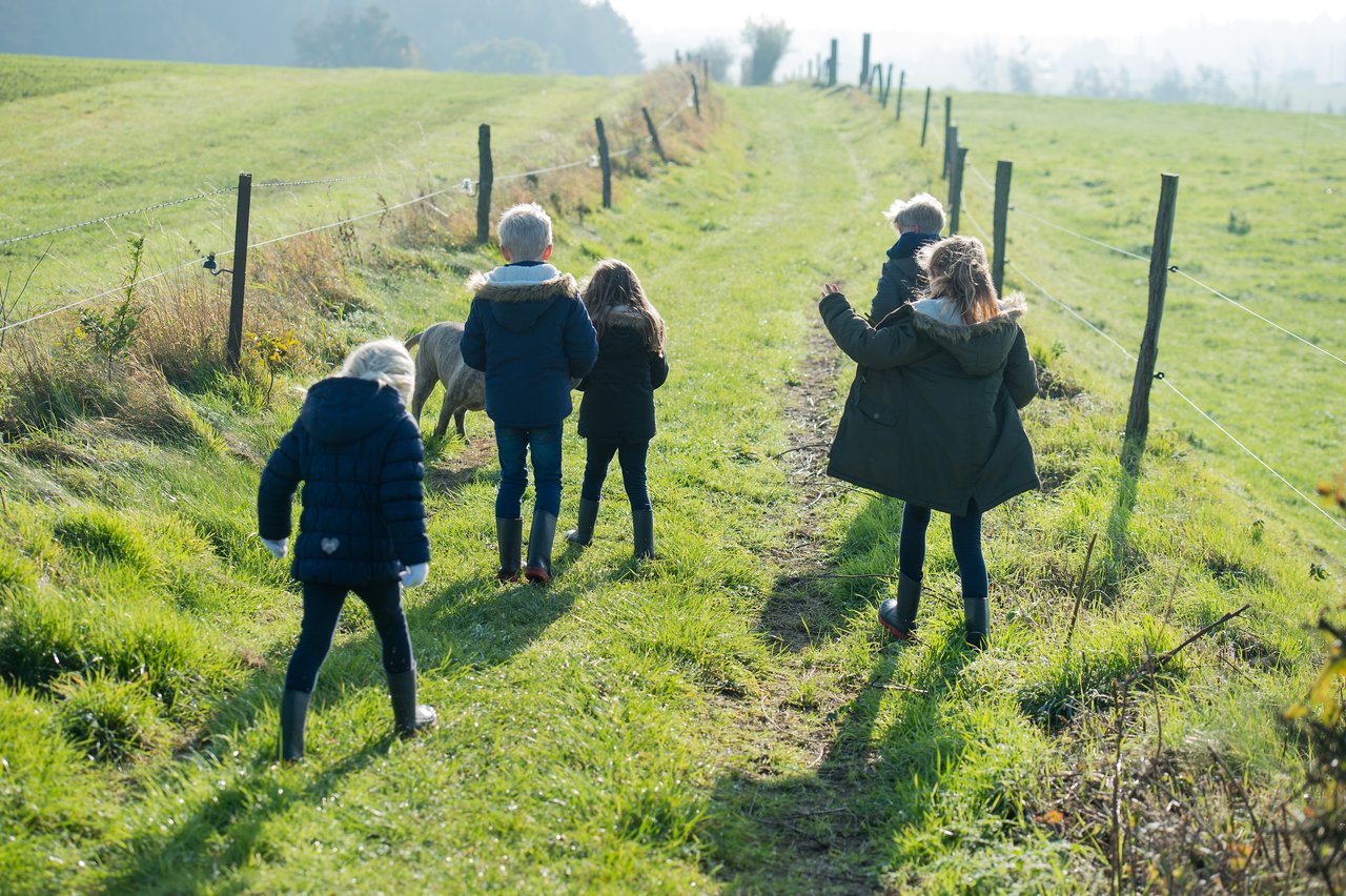 Five children in warm jackets walk along a grassy path with a small pony in a rural setting.