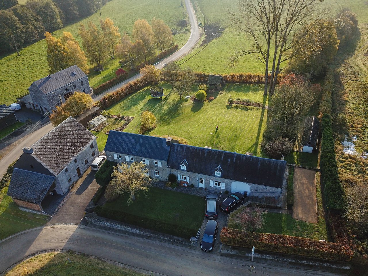 Aerial view of a countryside house with a large garden, parked cars, and a person standing on the lawn.