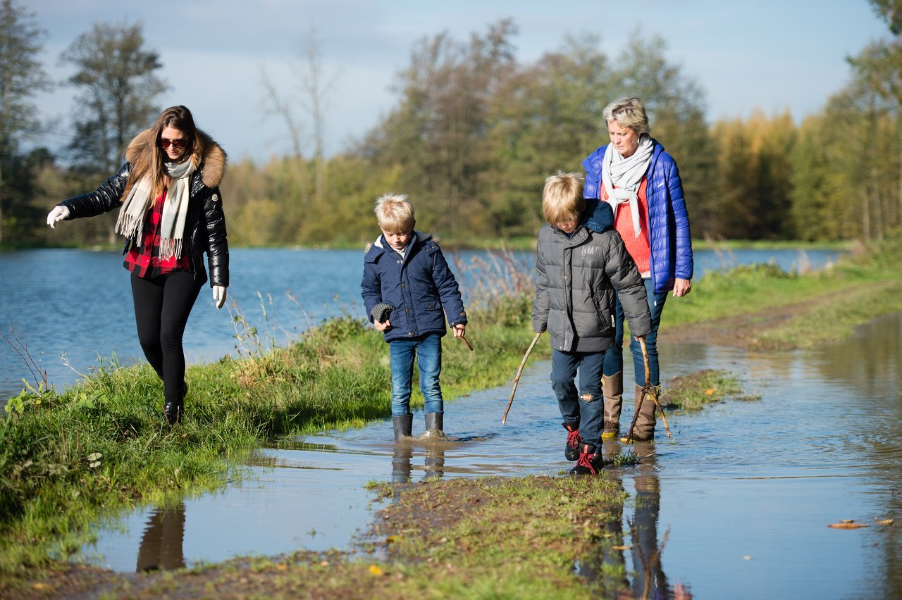 Two women and two children walk along a flooded path near a lake, carefully stepping through the water.