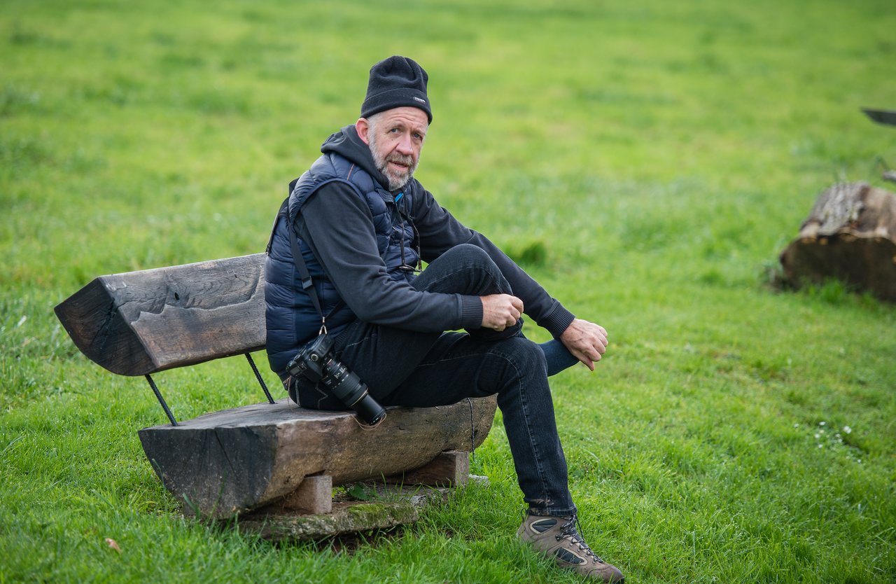 A man in outdoor clothing sits on a wooden bench in a grassy area, holding his leg.