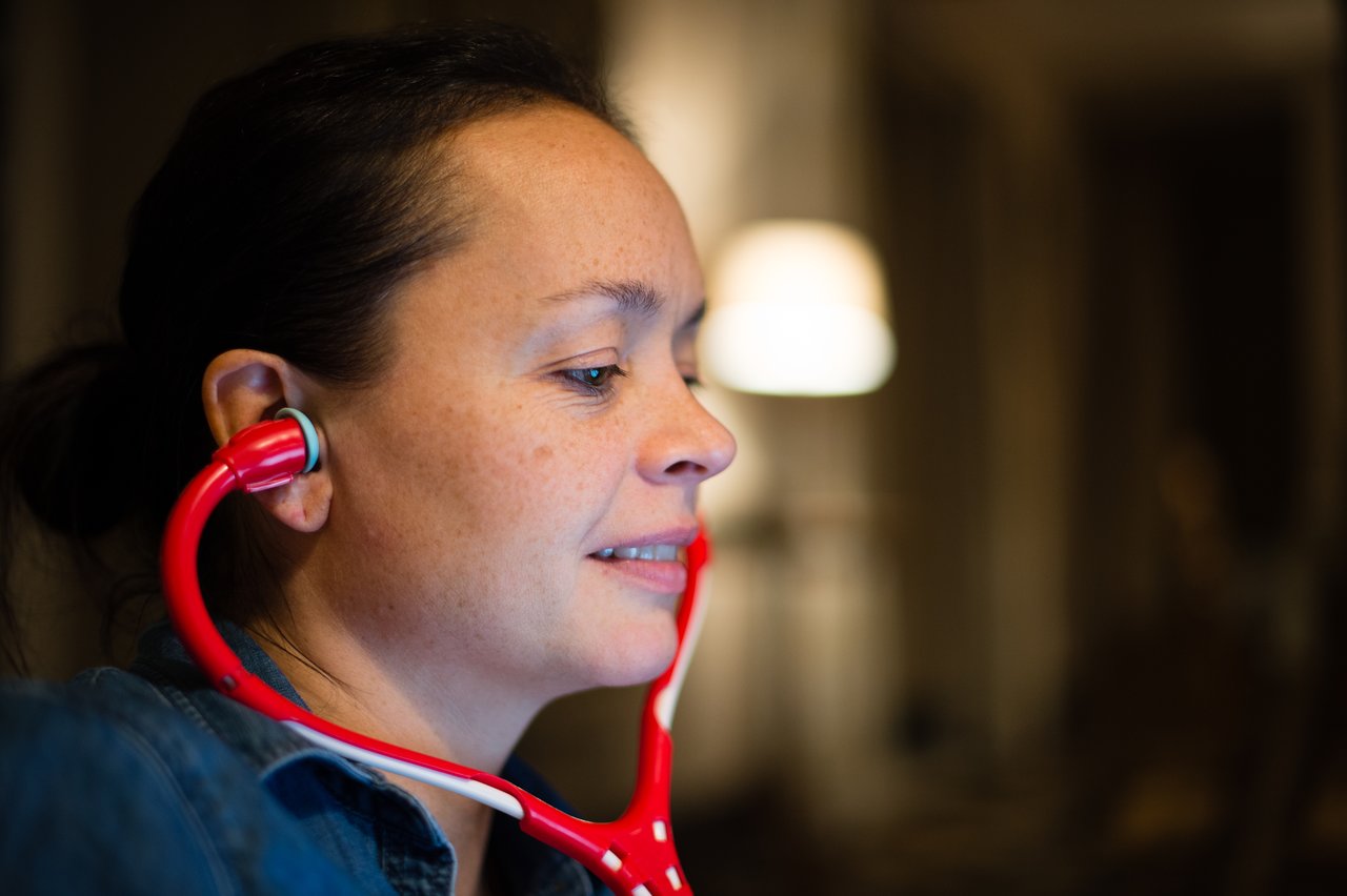 A woman wearing a red stethoscope listens through the earpieces while looking at something off-camera.