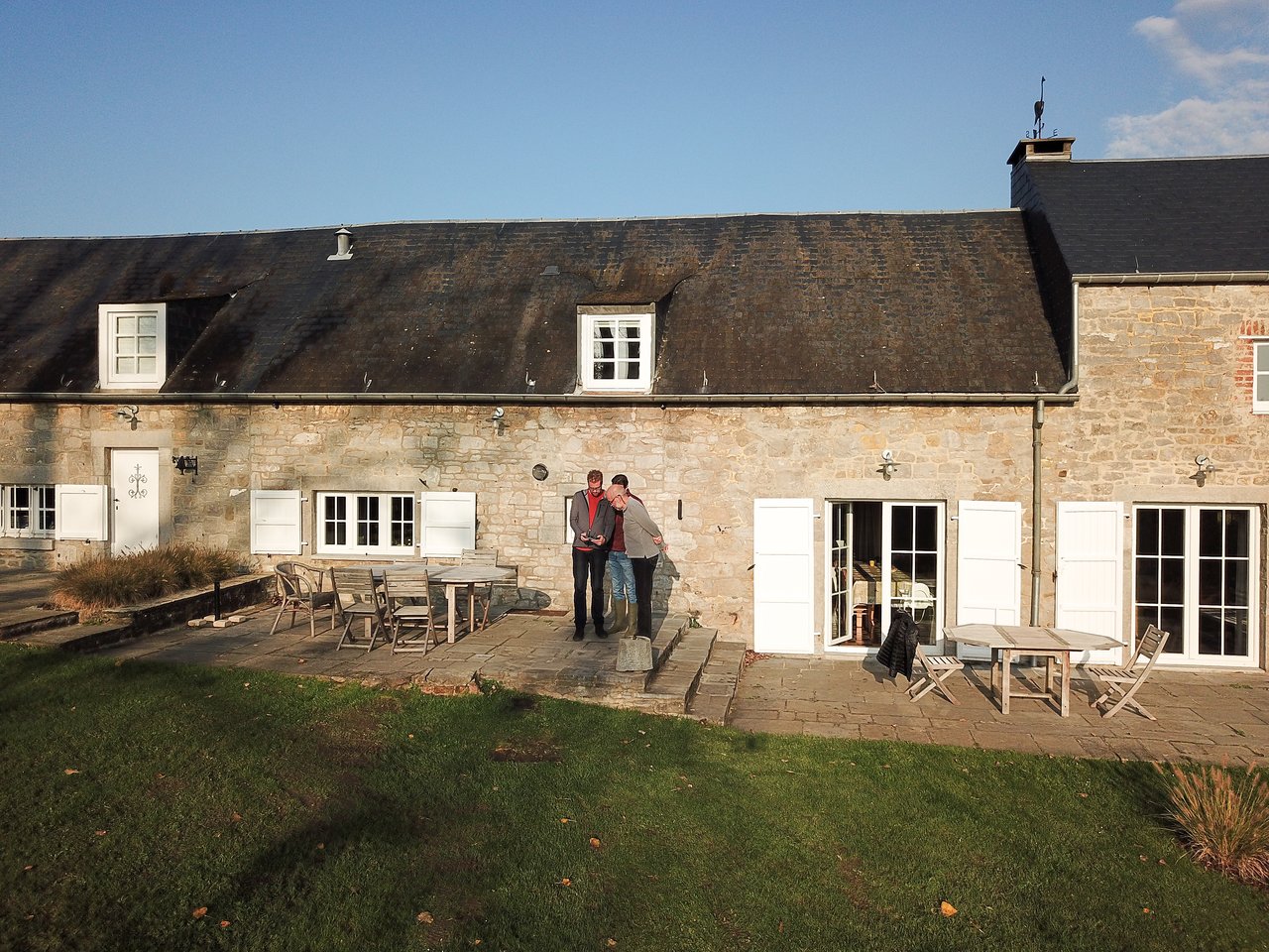 Two people stand on a stone patio, operating a drone in front of a rustic house with white shutters.