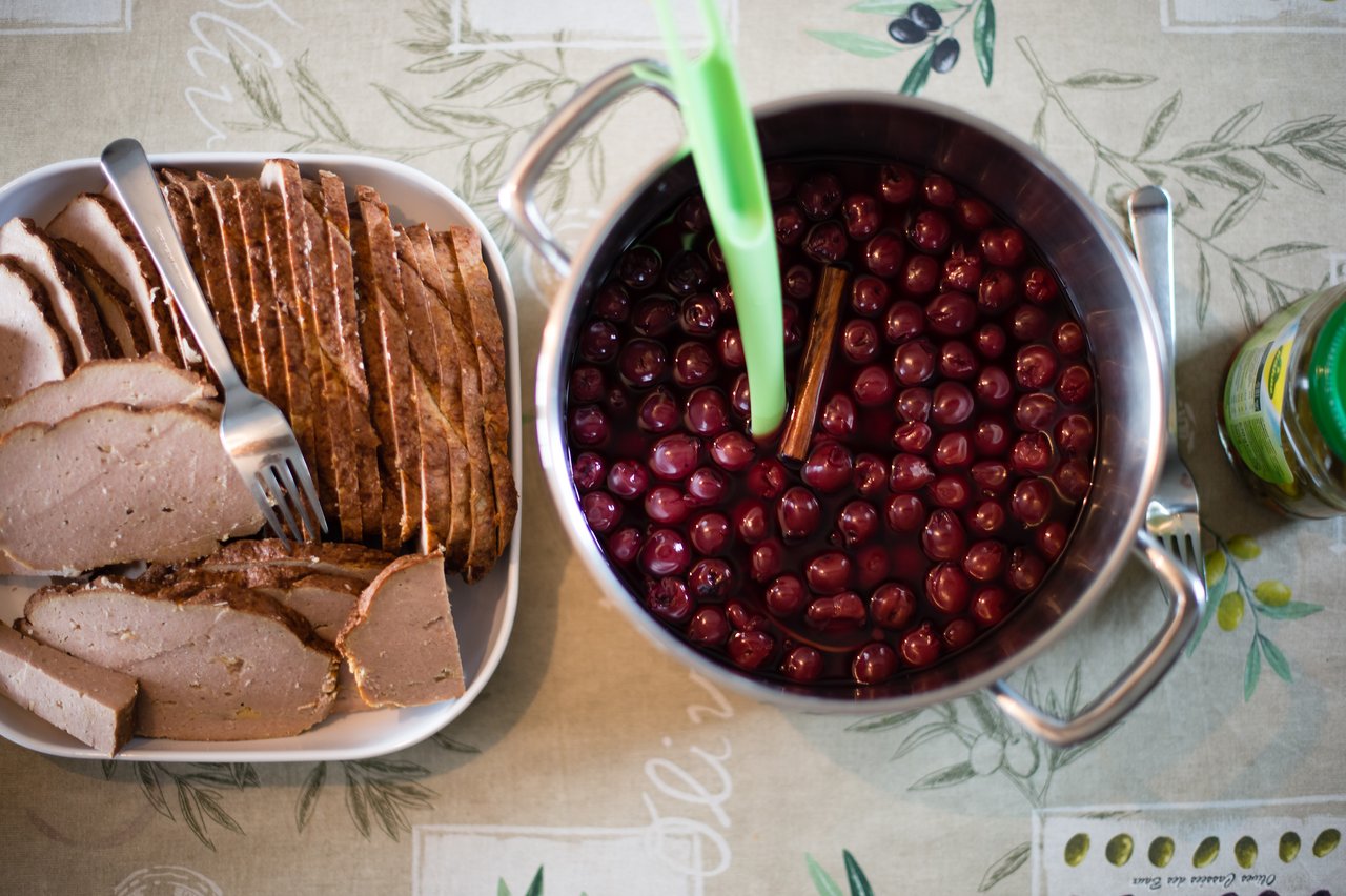 A plate of sliced meatloaf and a pot of cherries in syrup with a cinnamon stick.