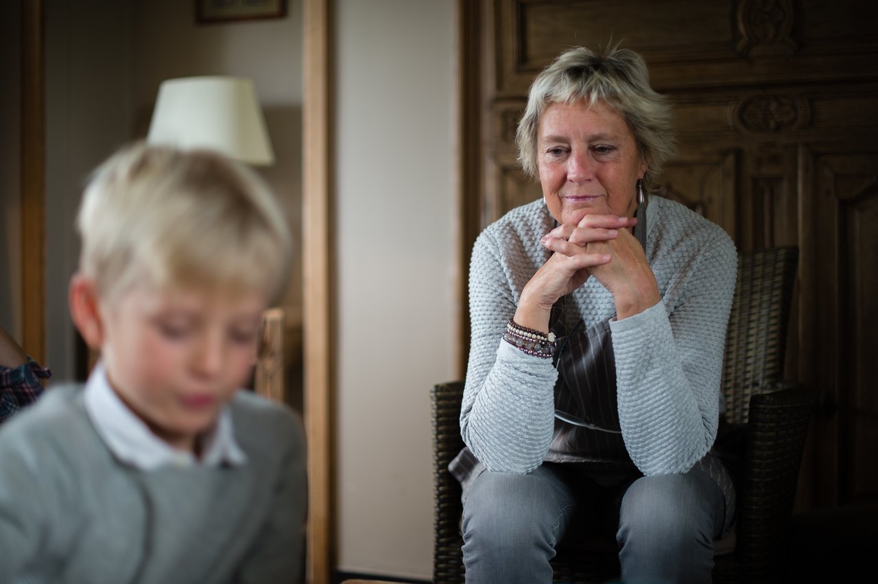 An older woman sits in a chair, resting her hands together, watching a young boy in the foreground.