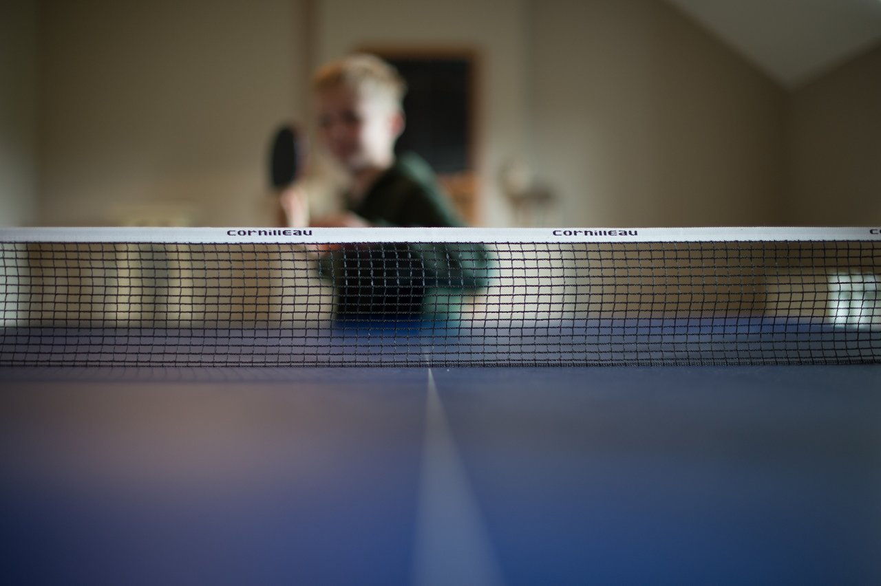 A person holding a paddle prepares to play ping pong, viewed from across the table with the net in focus.