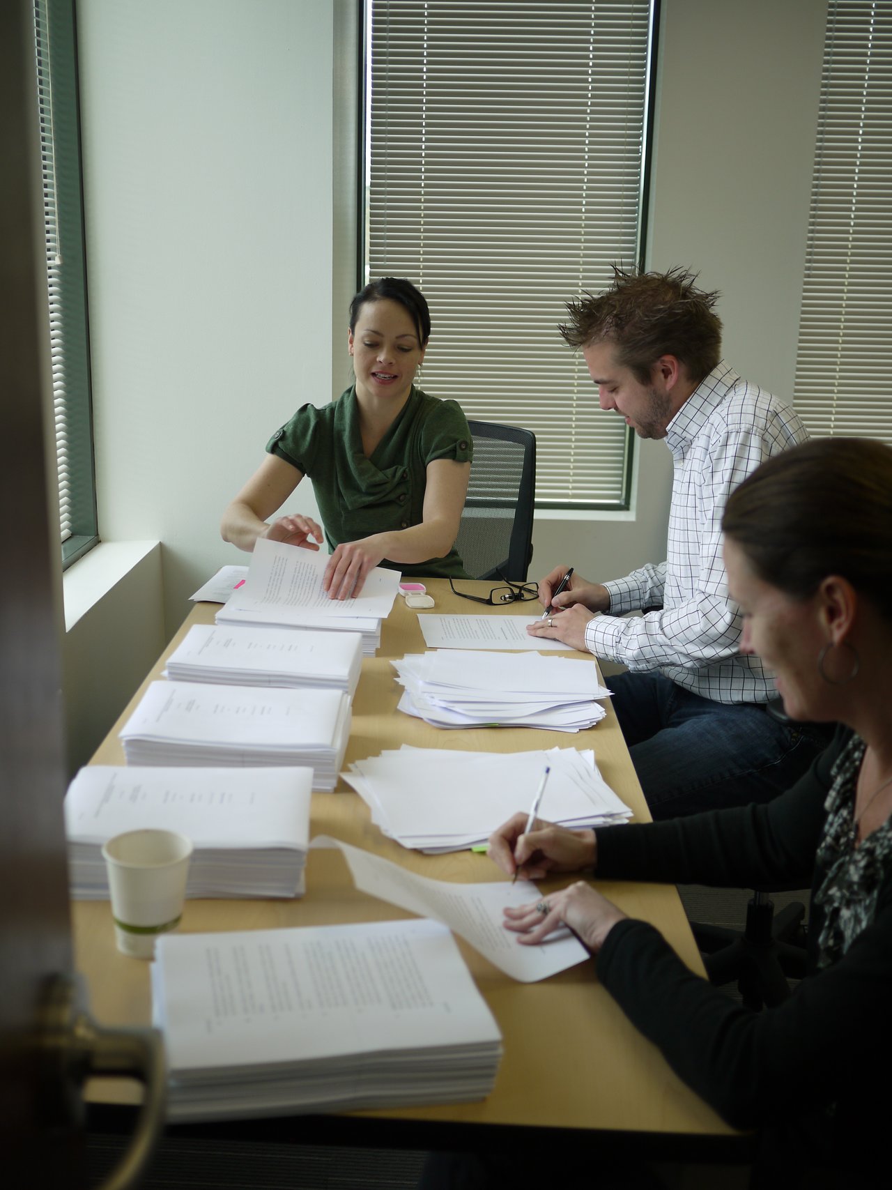 Three people sit at a table sorting and signing documents, with stacks of paper around them.
