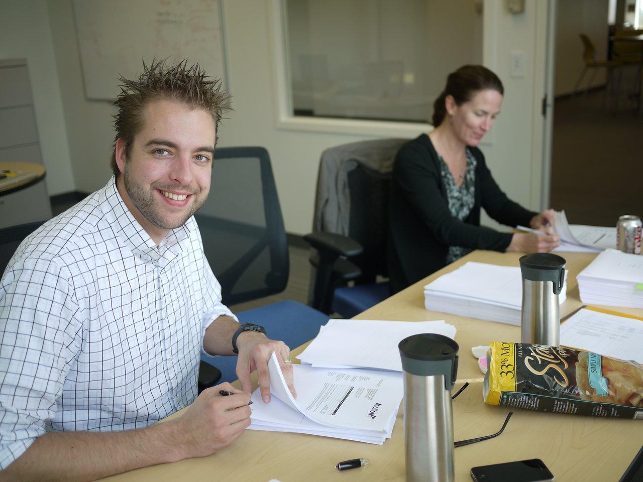 Two people sit at a table signing documents, with stacks of papers, coffee mugs, and snacks nearby.