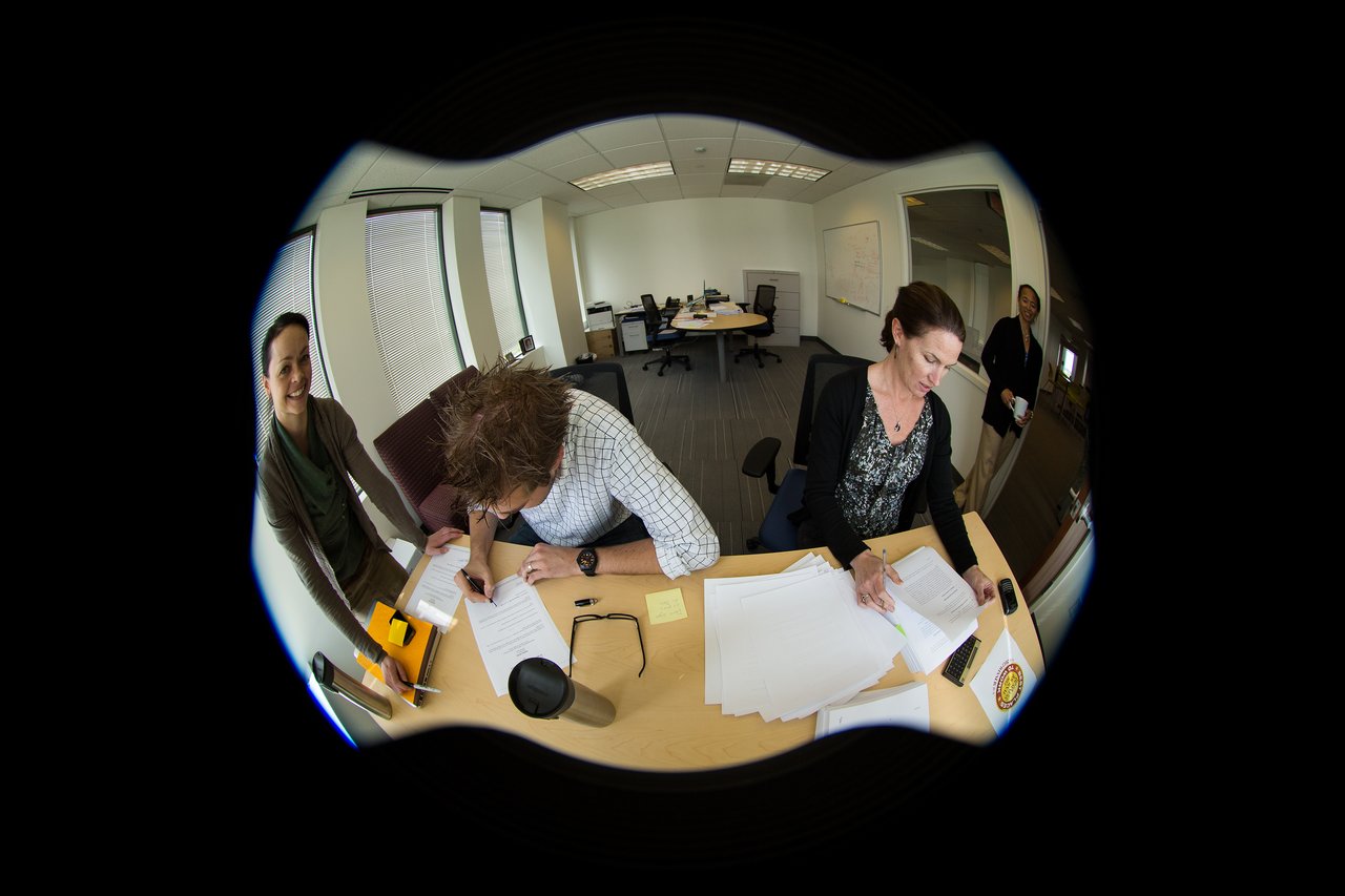 Two people sign documents at a desk while another person smiles nearby in an office setting.