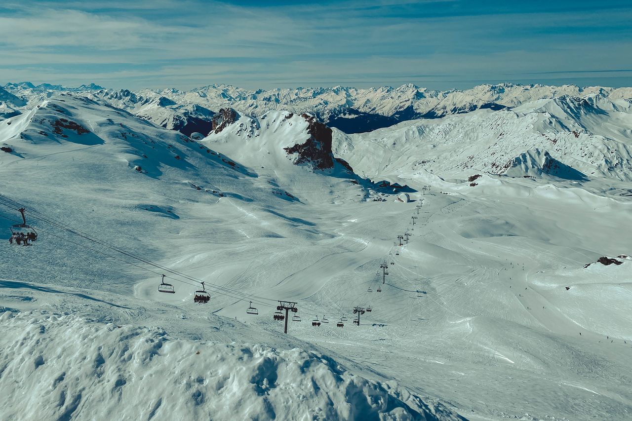 A long ski lift carries people up a snowy mountain with skiers and snowboarders visible on the slopes below.
