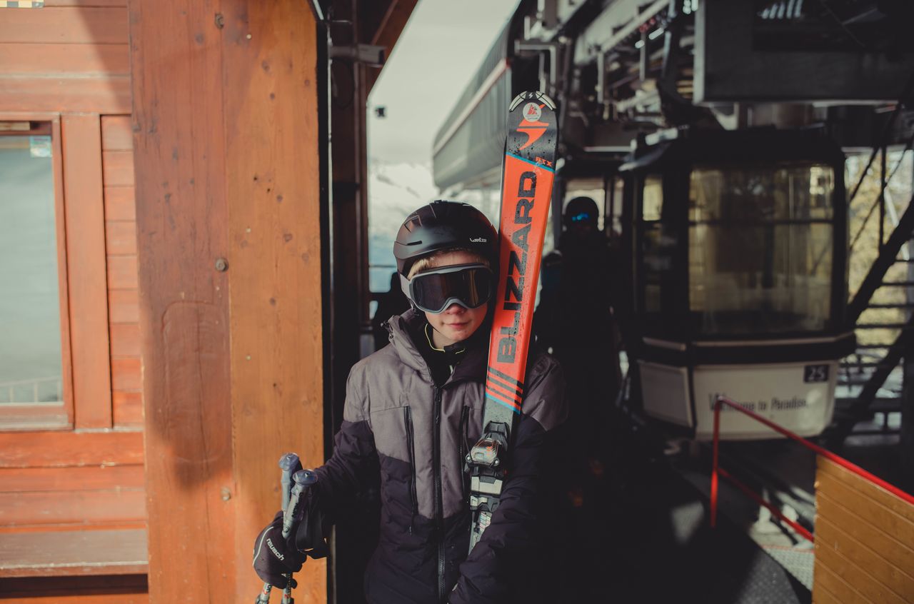 A young skier wearing a helmet and goggles holds skis and poles while standing near a ski lift station.