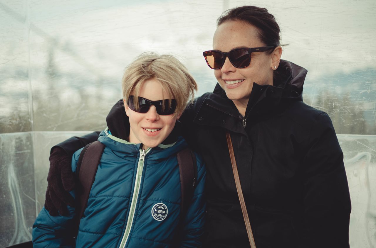 A woman and a child, both in sunglasses and winter jackets, smile while sitting closely together inside a gondola.
