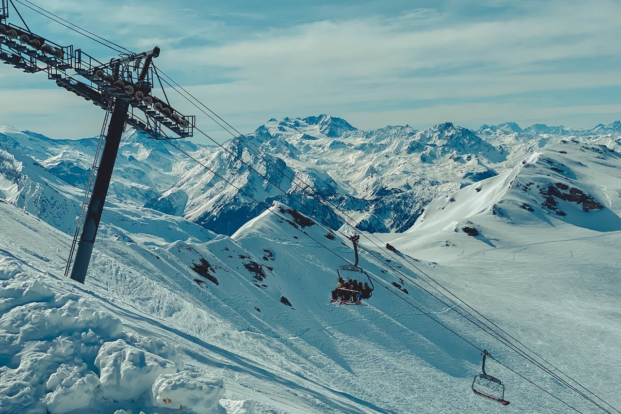 A ski lift carries a group of people up a snowy mountain with large peaks in the background.