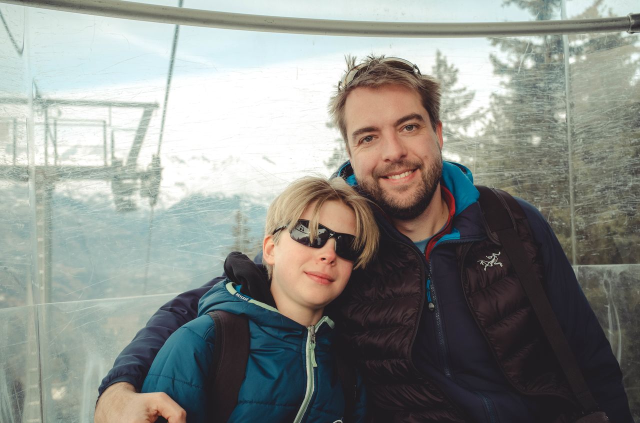 A man and a child, both in winter jackets, smile while sitting closely together inside a gondola.