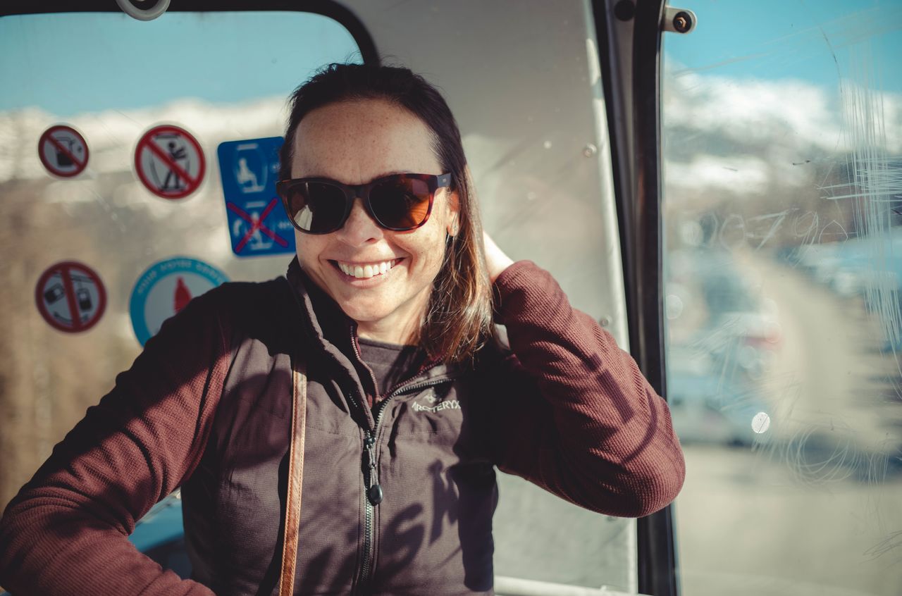 A woman wearing sunglasses and a jacket smiles while sitting inside a gondola lift.