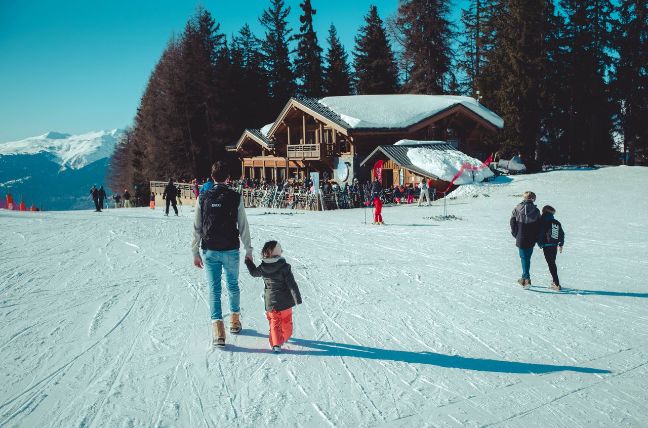 A man and a child walk hand in hand on a snowy slope near a mountain restaurant and bar.