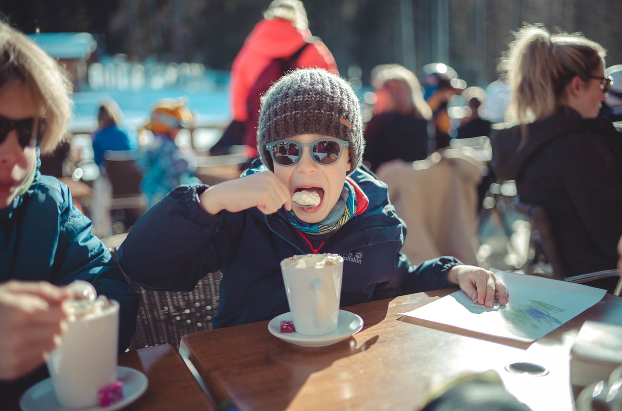 A child wearing a hat and sunglasses eats whipped cream from a spoon while sitting at an outdoor table.