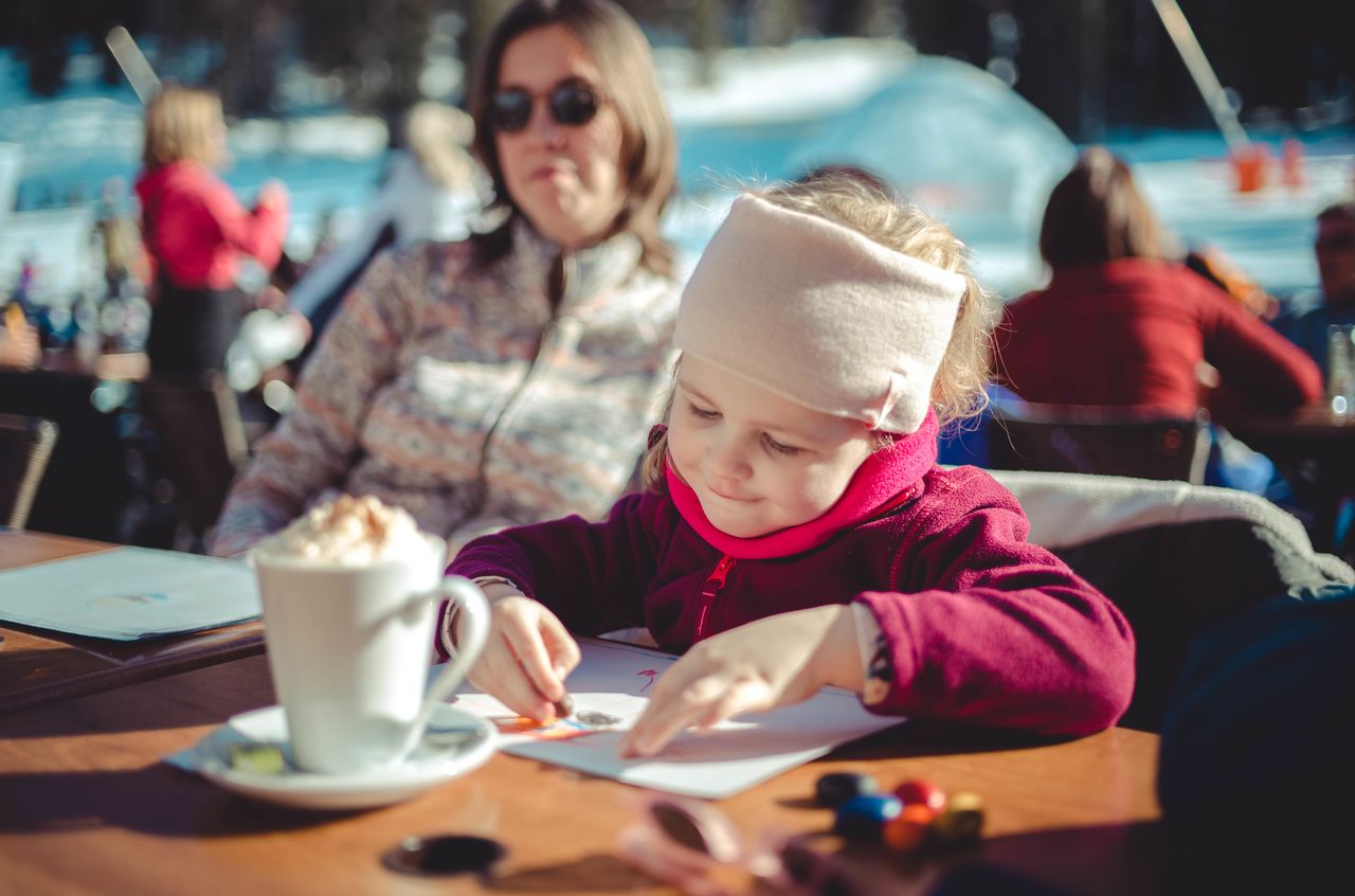 A young child in a red jacket colors on paper at an outdoor table.