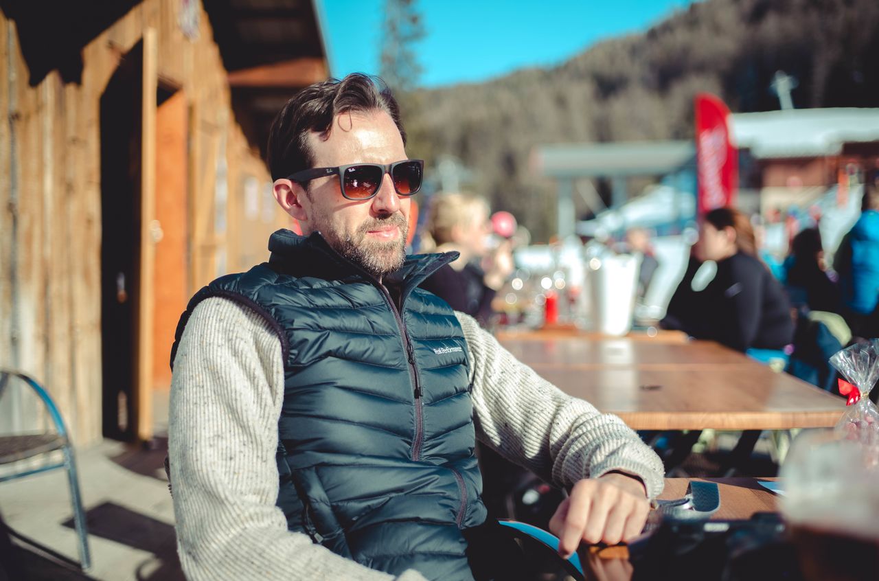 A man wearing sunglasses and a vest sits at an outdoor table, looking into the distance in sunlight.