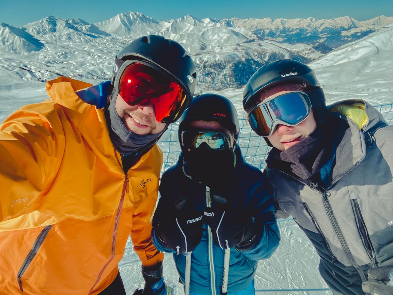 Three people in ski gear and helmets take a selfie on a snowy mountain, smiling behind their goggles.