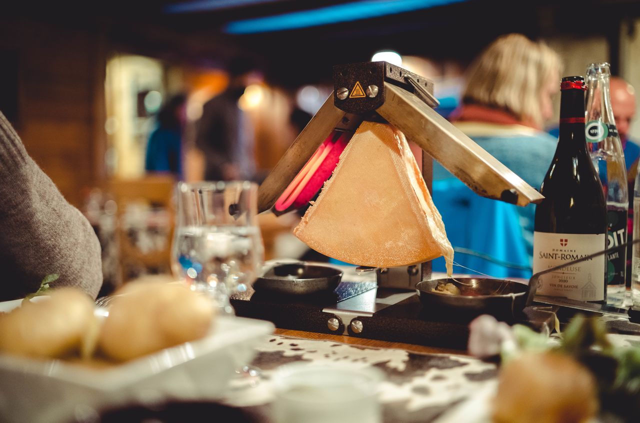 A wedge of cheese melts under a heating lamp, with potatoes, wine, and water on the table.
