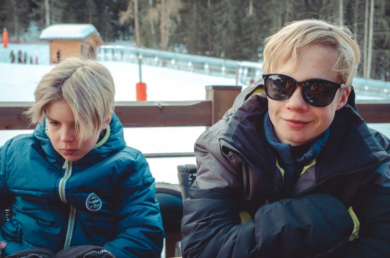 Two boys in winter jackets sit outdoors; one wears sunglasses and smiles, while the other looks down seriously.