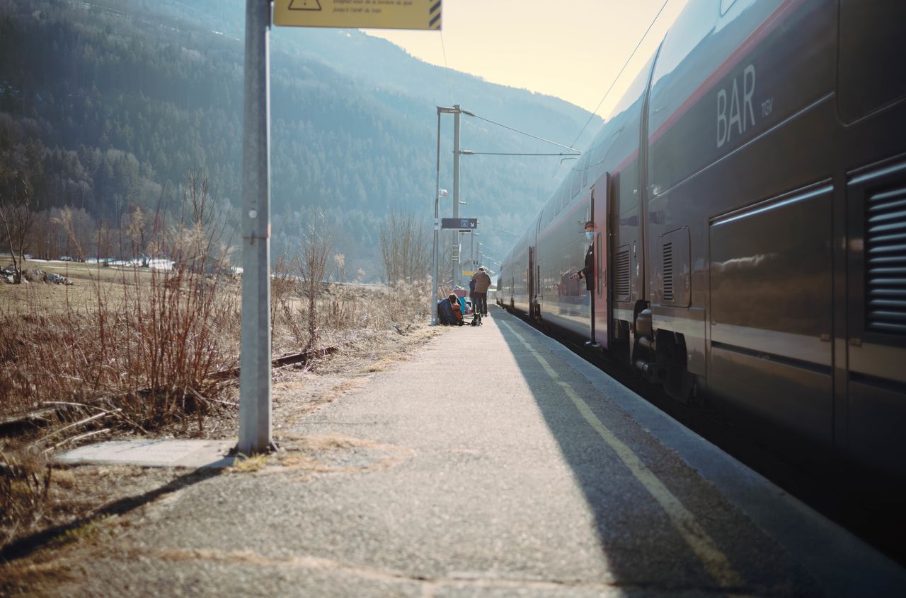 A train conductor leans out of the door while passengers wait on a rural train platform.