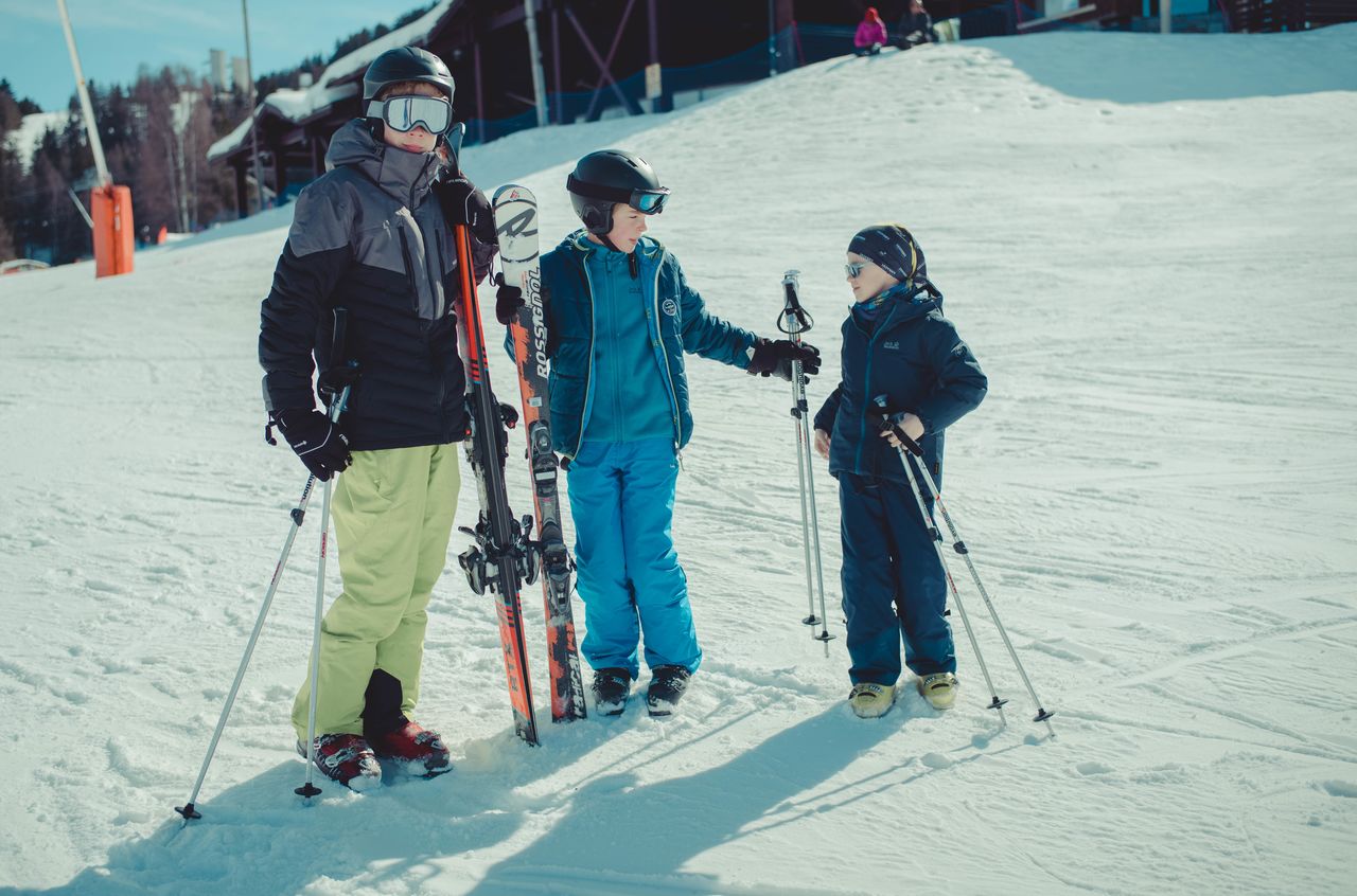 Three people in ski gear stand on a snowy slope, holding skis and poles while talking to each other.