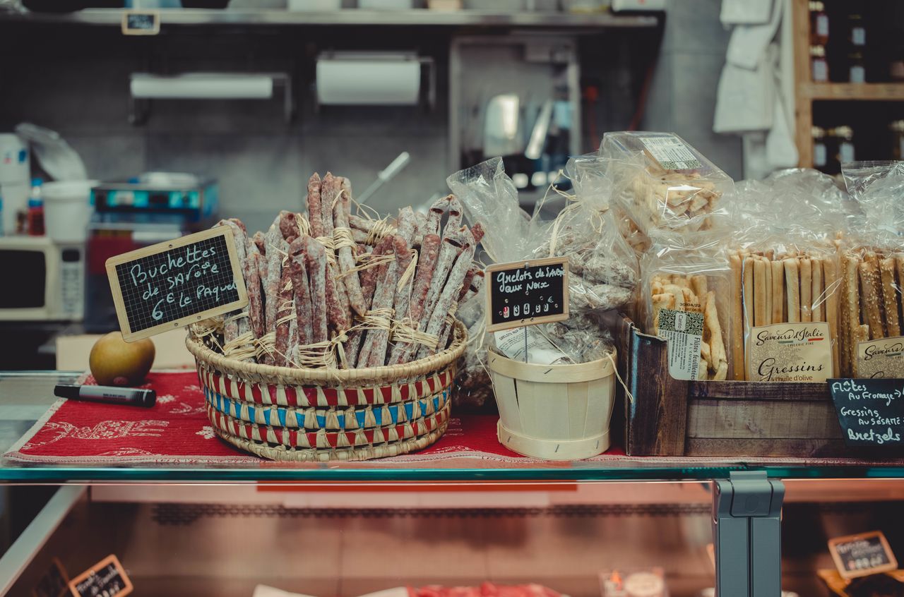 A market display with baskets of cured sausages, packaged breadsticks, and small chalkboard signs showing prices.