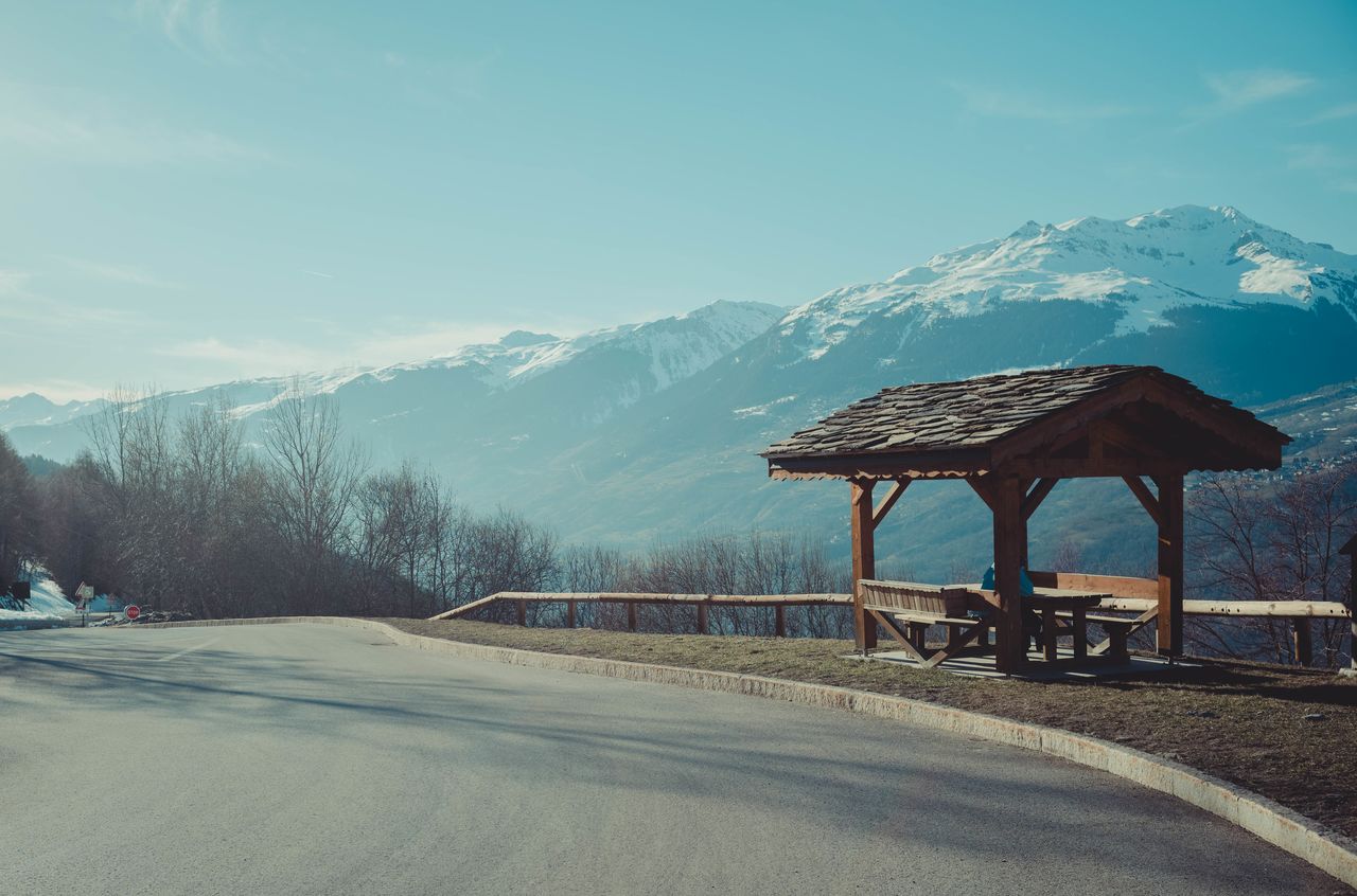 A wooden shelter at a roadside rest stop with snow-covered mountains in the background.