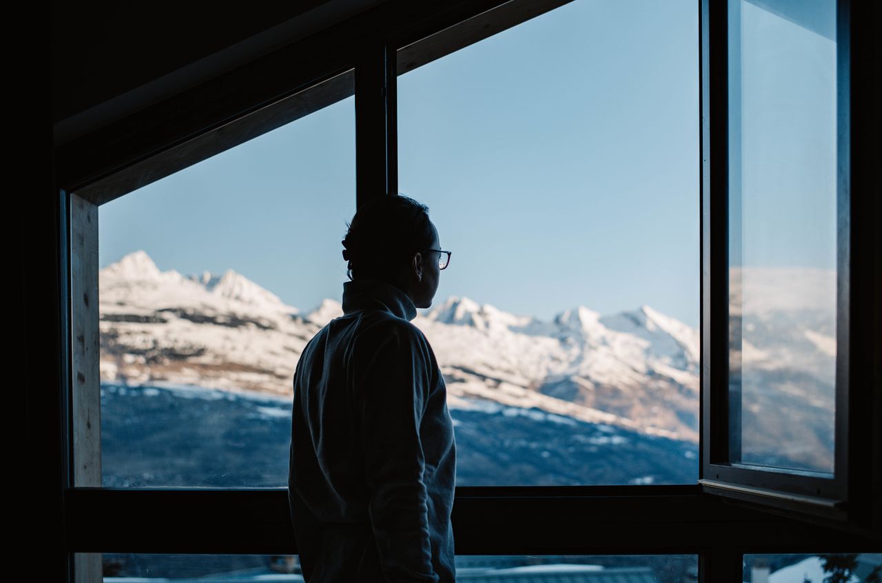 A woman wearing glasses looks out an open window at snow-covered mountains in the distance.