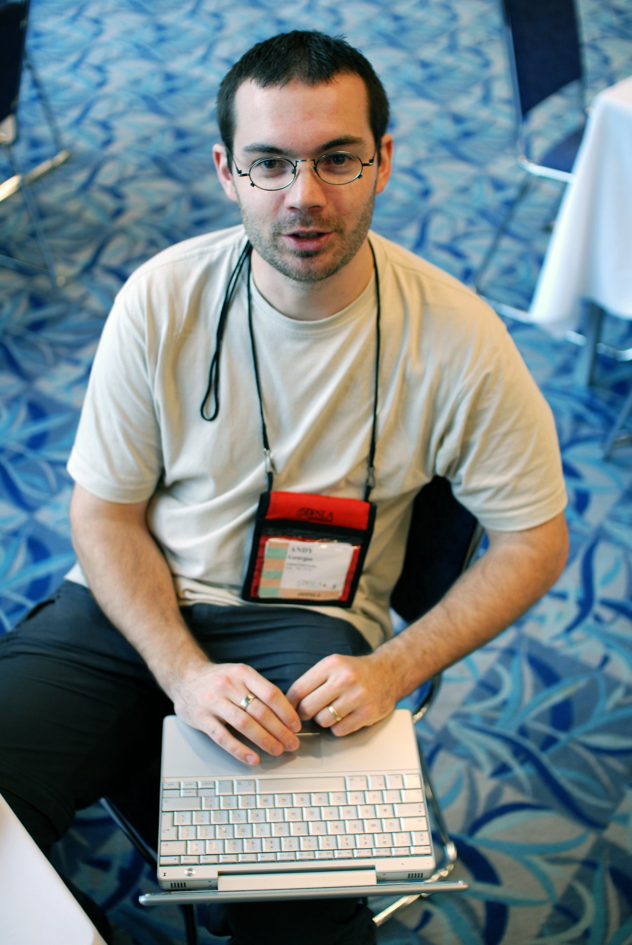 A man with glasses and a name badge sits and types on a laptop, looking up at the camera.