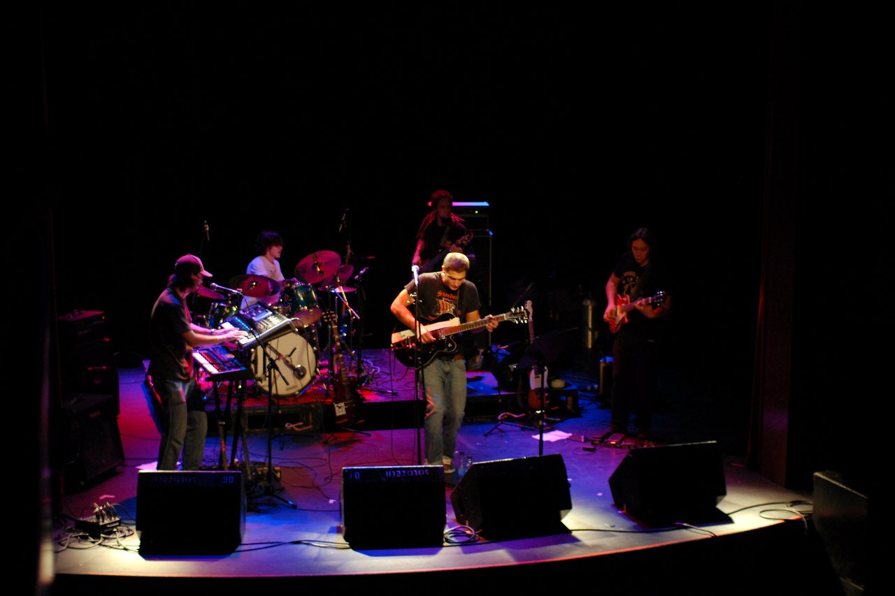 A five-member band performs on stage with guitars, keyboards, and drums under colorful stage lighting.