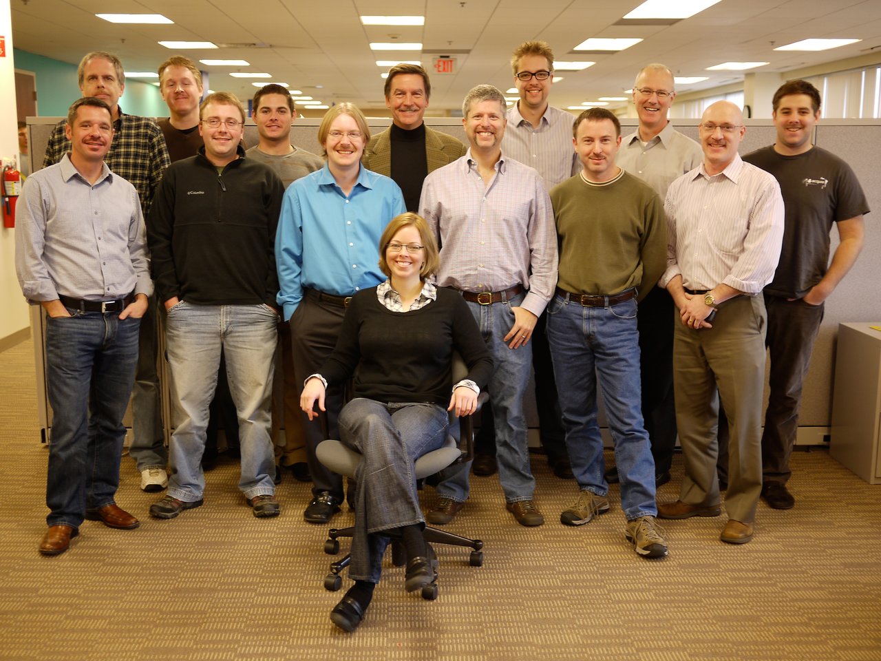 A group of fifteen Acquia team members pose in an office, with one person seated in front and others standing.