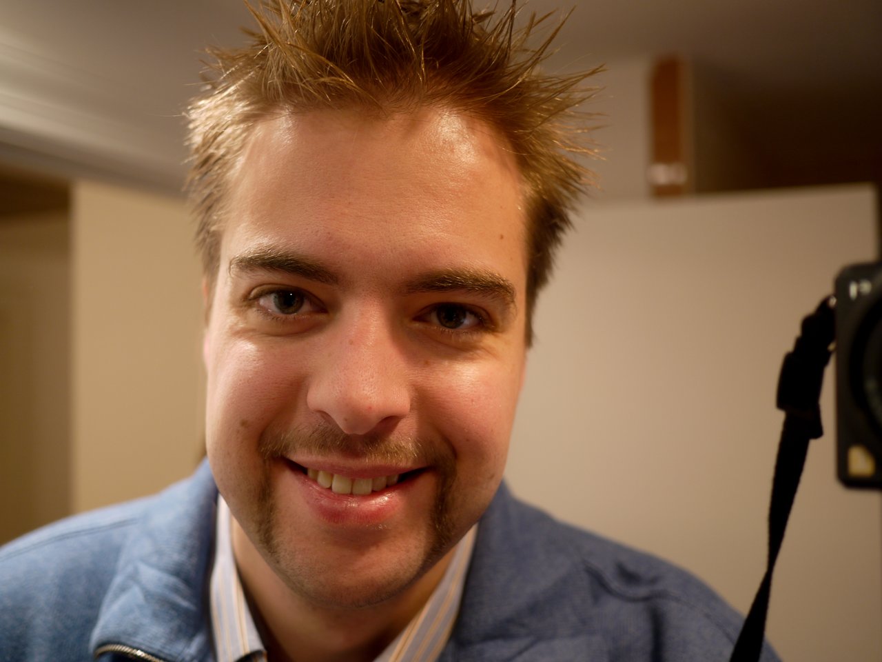 A man with short, spiky hair and a mustache smiles at the camera, wearing a blue jacket indoors.