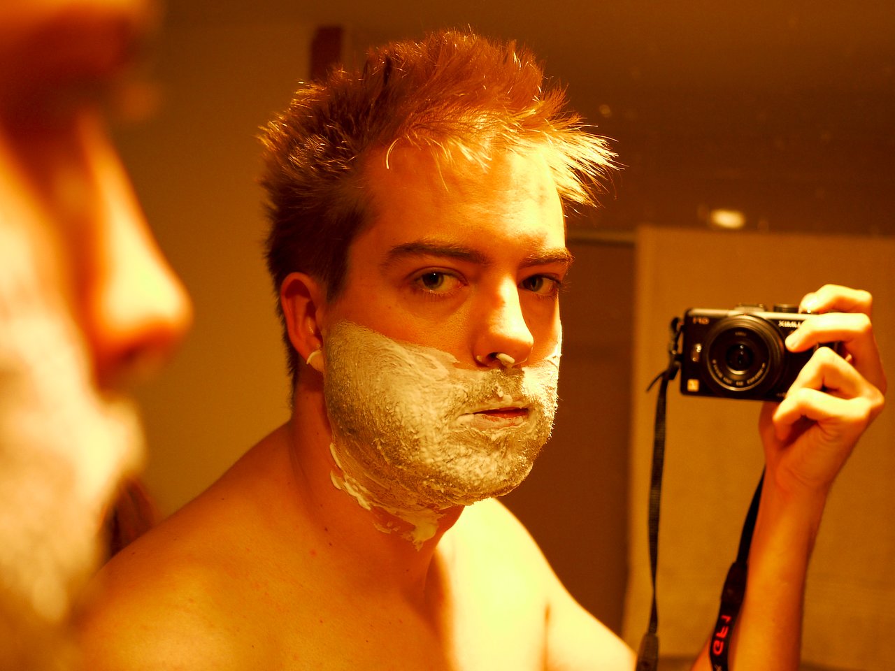 A man with shaving cream on his face takes a mirror selfie with a camera in a bathroom.
