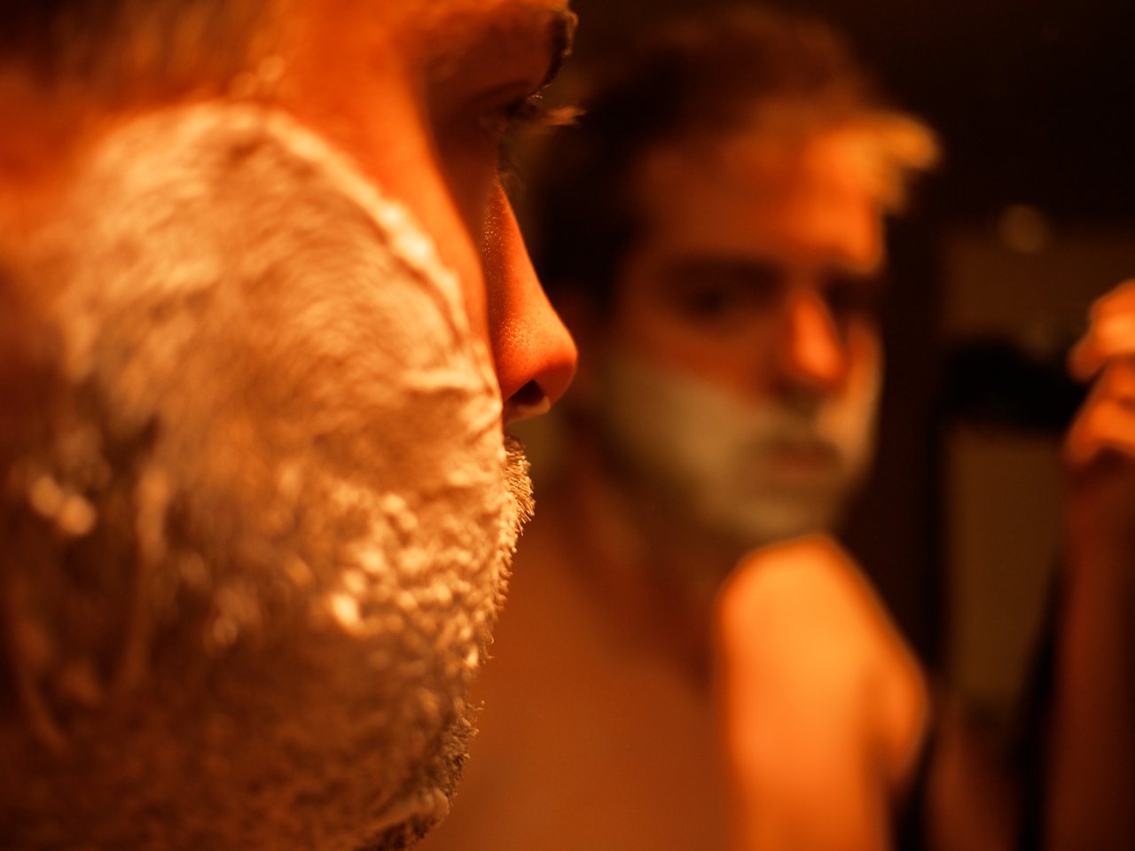 Close-up of a man's face with shaving cream, while his reflection in the mirror shows him shaving.